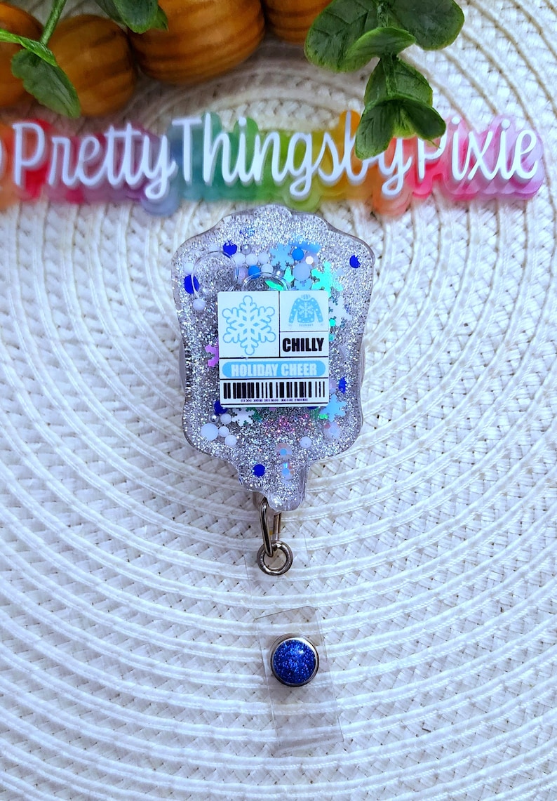 May include: A silver glitter badge holder with a blue snowflake and a white sweater with the text "CHILLY HOLIDAY CHEER". The badge holder has a blue glitter retractable badge reel.