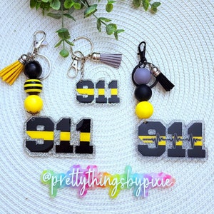 May include: Three keychains featuring the number 911 in black with a horizontal yellow stripe, set against a silver glitter background. Each includes a metal clasp, a decorative tassel, and round silicone beads in yellow, black, or gray.