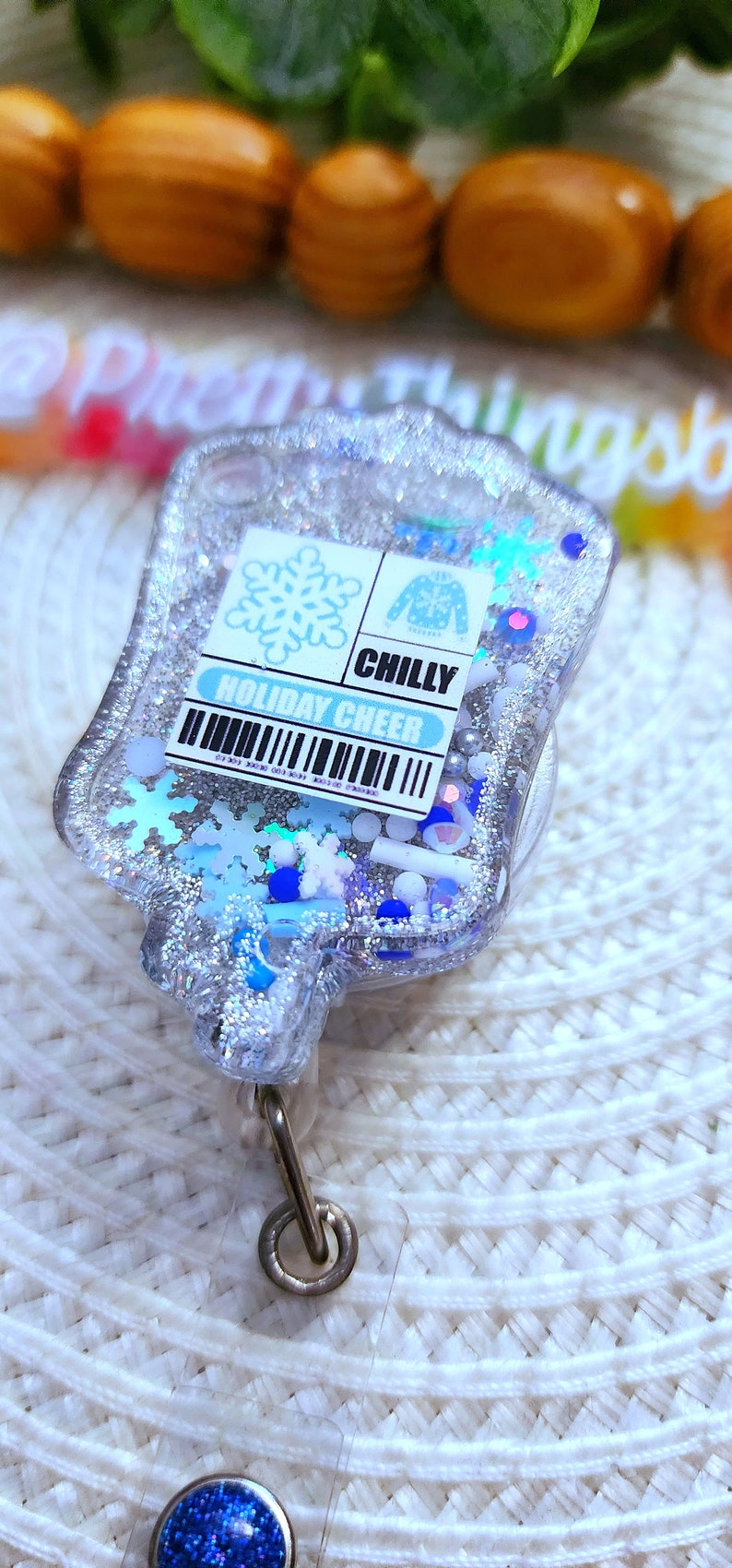 May include: A silver glitter badge holder with a blue and white snowflake design. The badge holder has a clear plastic front and a silver metal clip. The text "Holiday Cheer" and "Chilly" are printed on the badge holder.