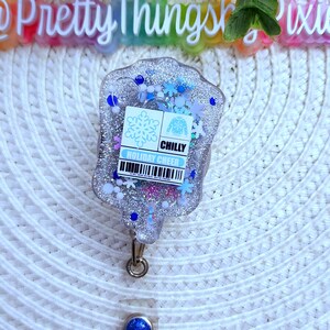 May include: A clear, silver glitter badge holder with a blue snowflake and sweater design. The text "CHILLY HOLIDAY CHEER" is printed on the badge holder.
