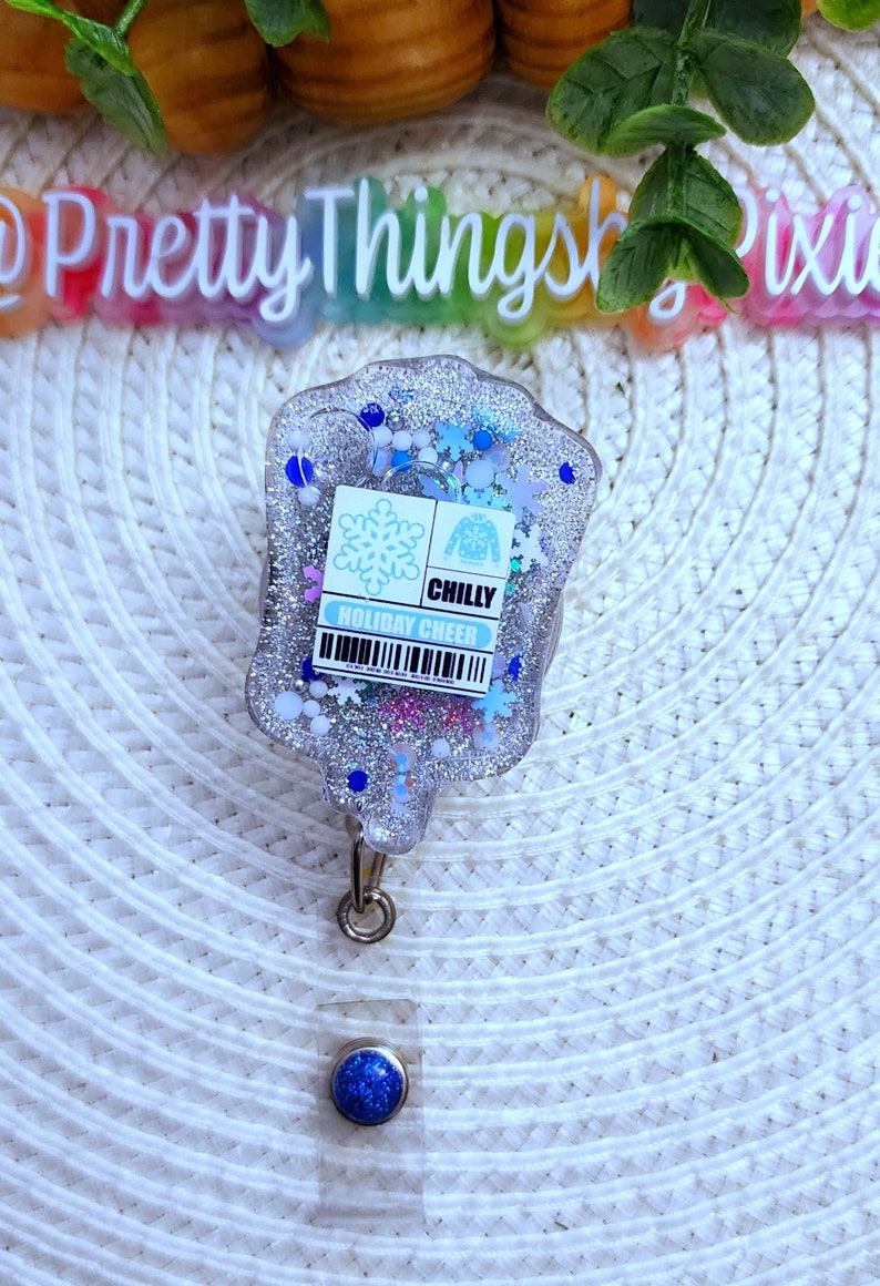 May include: A clear, silver glitter badge holder with a blue snowflake and sweater design. The text "CHILLY HOLIDAY CHEER" is printed on the badge holder.