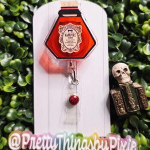 May include: A red, hexagonal badge reel shaped like a potion bottle, labeled "Vampire Venom." The reel has a clear plastic clip and a glittery red accent. The background is a white surface with green foliage.