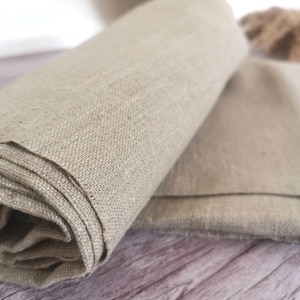 Puede incluir: Tela de lino beige enrollada, con un tejido texturizado. La tela está doblada sobre otro trozo del mismo material, con los bordes visibles. El fondo es una superficie de madera.