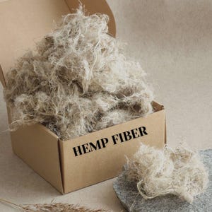 Può includere: Una scatola di cartone piena di fibra di canapa marrone chiaro. La scatola è aperta, rivelando una grande quantità di materiale fibroso. Le parole "HEMP FIBER" sono stampate in nero sulla parte anteriore della scatola. Un piccolo mucchio di fibra di canapa è su una pietra grigia.