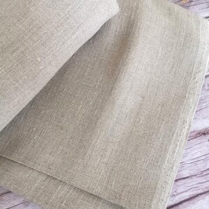 Close-up of folded, light beige linen fabric. The fabric has a textured appearance with visible weave details. The folds create shadows, highlighting the material's natural texture. The neutral color suggests a versatile textile for various uses.