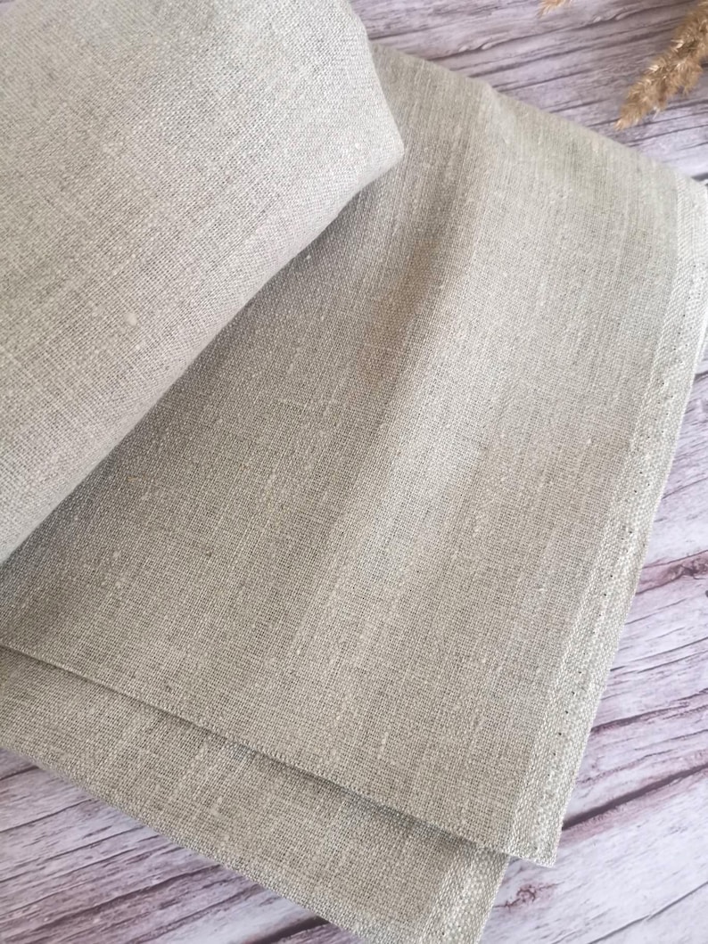 Close-up of folded, light beige linen fabric. The fabric has a textured appearance with visible weave details. The folds create shadows, highlighting the material's natural texture. The neutral color suggests a versatile textile for various uses.