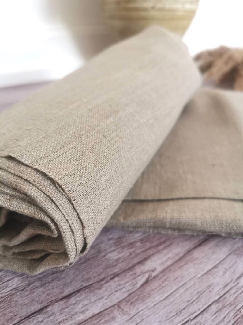 A rolled-up, beige linen fabric with a textured weave. The fabric is folded over another piece of the same material, with the edges visible. The background is a wooden surface.