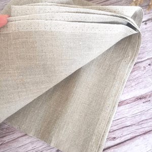 K&ouml;nnte beinhalten: Gefaltete Leinen-Serviette in einem neutralen Beige. Der Stoff hat eine strukturierte Optik und ein sichtbares Gewebe. Die Kanten sind sauber ges&auml;umt, und die Serviette ist teilweise entfaltet, wodurch ihre Schichten sichtbar werden. Der Hintergrund ist eine Holzoberfl&auml;che.