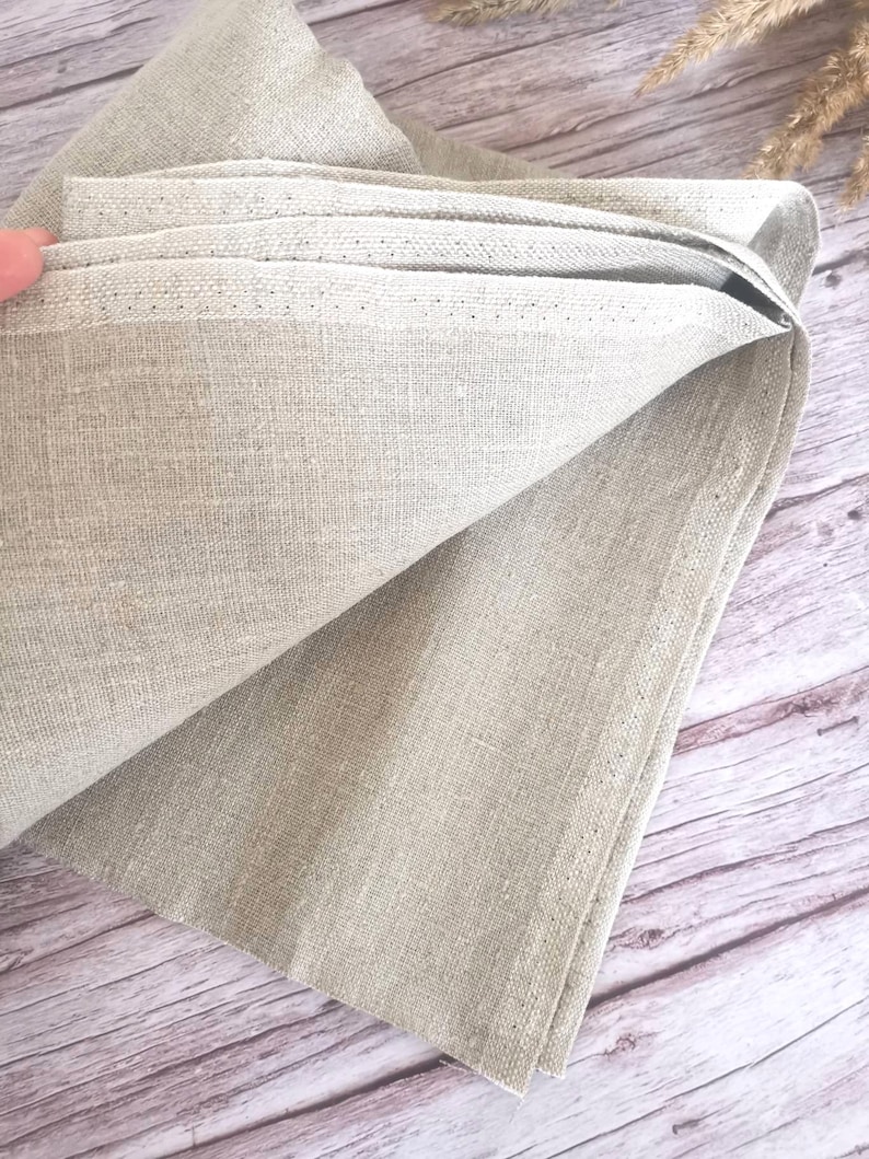 K&ouml;nnte beinhalten: Gefaltete Leinen-Serviette in einem neutralen Beige. Der Stoff hat eine strukturierte Optik und ein sichtbares Gewebe. Die Kanten sind sauber ges&auml;umt, und die Serviette ist teilweise entfaltet, wodurch ihre Schichten sichtbar werden. Der Hintergrund ist eine Holzoberfl&auml;che.
