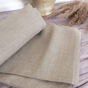 K&ouml;nnte beinhalten: Gefalteter beigefarbener Leinenstoff, wahrscheinlich f&uuml;r eine Tischdecke oder ein &auml;hnliches Textil. Der Stoff hat ein nat&uuml;rliches, leicht strukturiertes Aussehen. Das Bild enth&auml;lt auch eine dekorative Vase und getrocknetes Ziergras, was auf eine rustikale oder l&auml;ndliche &Auml;sthetik hindeutet.