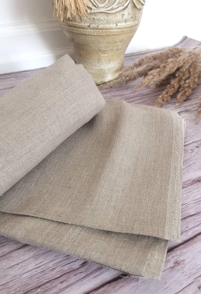 K&ouml;nnte beinhalten: Gefalteter beigefarbener Leinenstoff, wahrscheinlich f&uuml;r eine Tischdecke oder ein &auml;hnliches Textil. Der Stoff hat ein nat&uuml;rliches, leicht strukturiertes Aussehen. Das Bild enth&auml;lt auch eine dekorative Vase und getrocknetes Ziergras, was auf eine rustikale oder l&auml;ndliche &Auml;sthetik hindeutet.