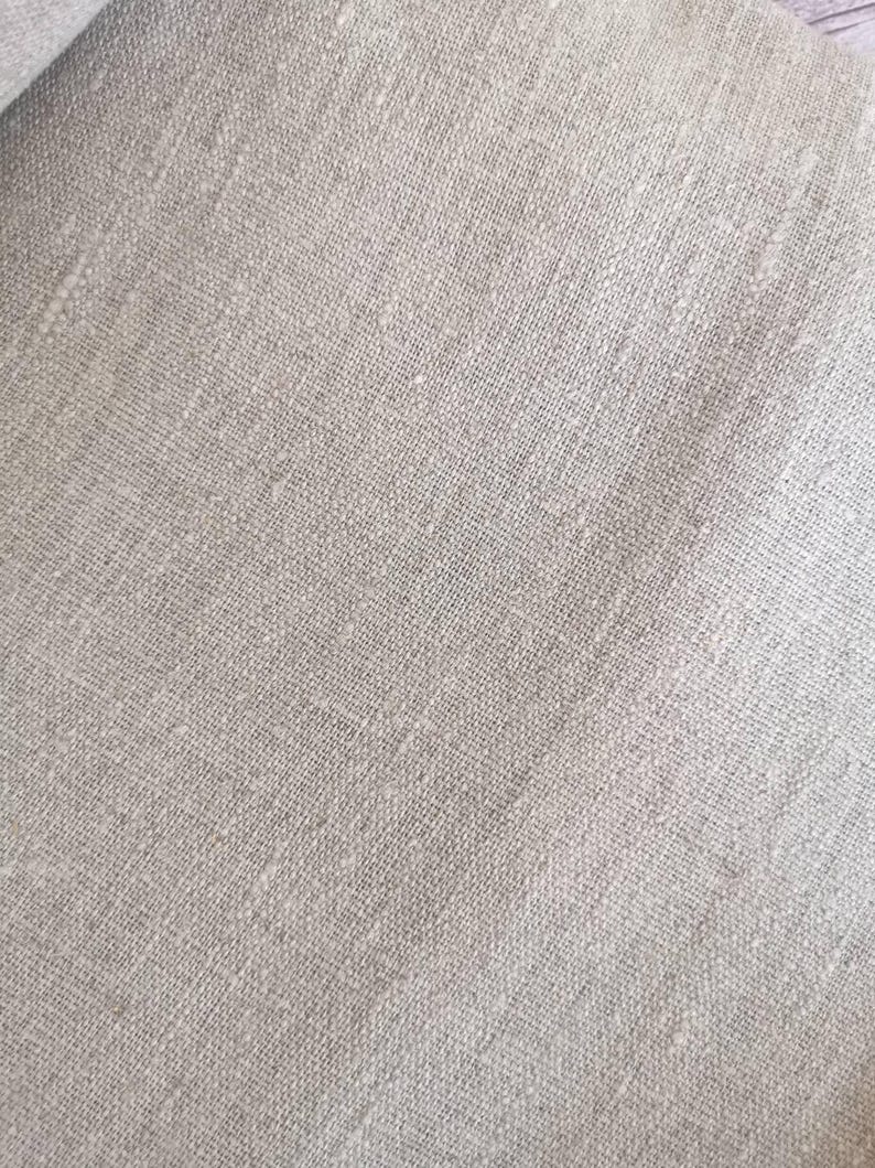 K&ouml;nnte beinhalten: Nahaufnahme eines hellbeigen Leinenstoffs. Der Stoff hat ein strukturiertes Aussehen mit sichtbarem Gewebe und subtilen Farbvariationen. Das Material wirkt weich und leicht zerknittert, was auf eine nat&uuml;rliche Textilfaser hindeutet.