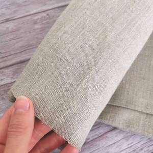 K&ouml;nnte beinhalten: Nahaufnahme eines aufgerollten, hellgrauen Leinenstoffs. Der Stoff hat eine strukturierte Optik und wird von einer Hand gehalten, die den Rand und das Gewebe des Materials zeigt. Der Hintergrund ist eine helle Holzoberfl&auml;che.