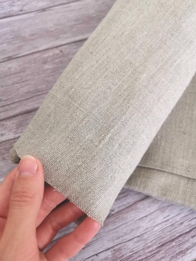 K&ouml;nnte beinhalten: Nahaufnahme eines aufgerollten, hellgrauen Leinenstoffs. Der Stoff hat eine strukturierte Optik und wird von einer Hand gehalten, die den Rand und das Gewebe des Materials zeigt. Der Hintergrund ist eine helle Holzoberfl&auml;che.