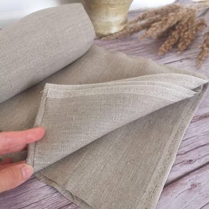 A folded, light gray linen napkin with a rolled-up section in the background. The fabric has a textured appearance. The napkin is on a wooden surface, with a decorative vase and dried plants in the background.