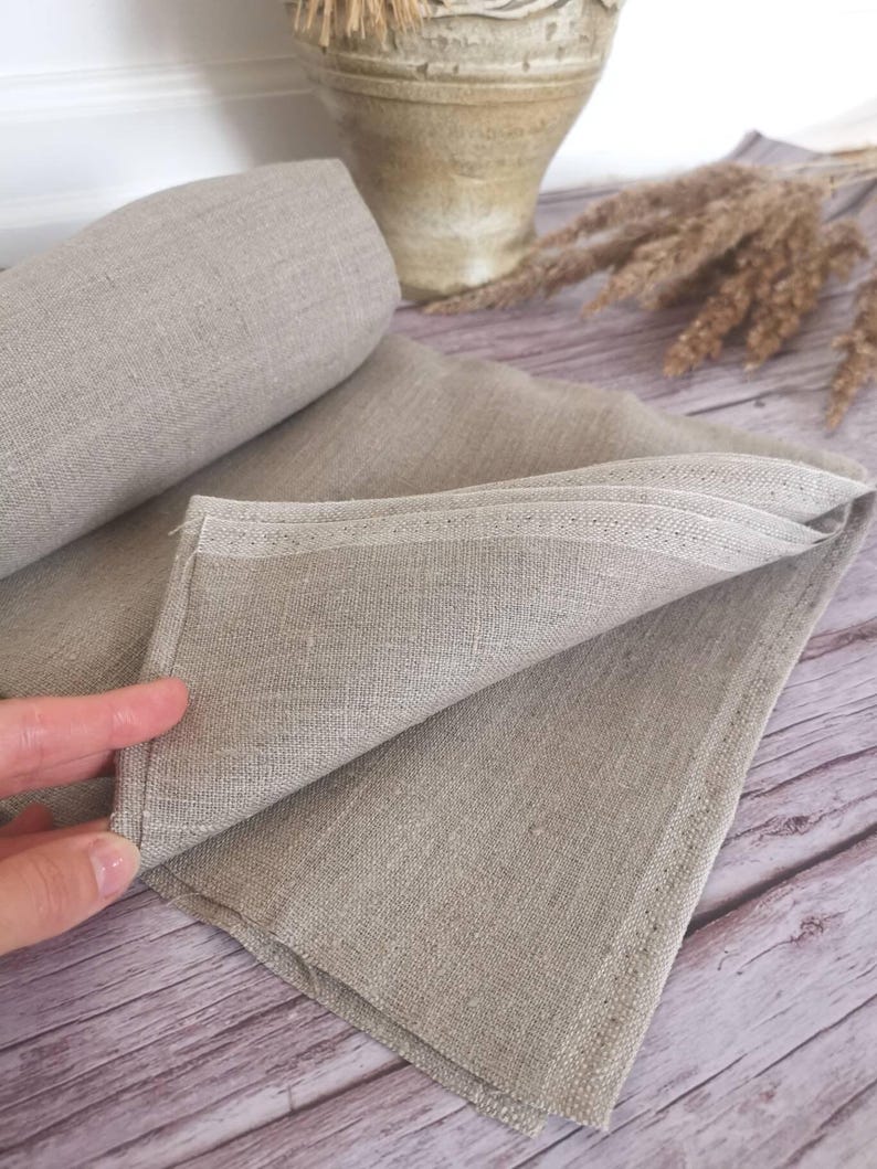 A folded, light gray linen napkin with a rolled-up section in the background. The fabric has a textured appearance. The napkin is on a wooden surface, with a decorative vase and dried plants in the background.