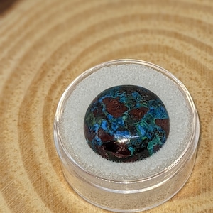 May include: A close-up of a round, blue, green, and brown gemstone cabochon. The gemstone is set in a clear plastic container.