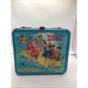 May include: Vintage blue metal lunchbox featuring a colorful illustration from "Bedknobs and Broomsticks." The artwork depicts characters on a bed, with the film's title prominently displayed. The lunchbox has a brown handle and clasp.