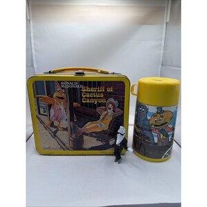 May include: A vintage yellow Ronald McDonald "Sheriff of Cactus Canyon" lunchbox and matching thermos. The lunchbox features a colorful graphic of Ronald McDonald and friends. The thermos has a yellow lid and a cartoon illustration.