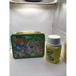 May include: Vintage Cabbage Patch Kids lunchbox and thermos set. The lunchbox is a rectangular metal container with a yellow handle and green trim. The thermos is yellow with a white lid and a Cabbage Patch Kids graphic.