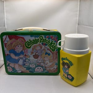May include: Vintage Cabbage Patch Kids lunchbox and thermos set. The lunchbox is green with a colorful illustration of Cabbage Patch Kids. The thermos is yellow with a white lid and handle, also featuring Cabbage Patch Kids graphics.