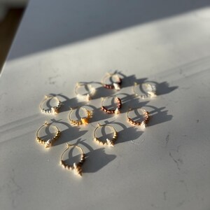 May include: A selection of gold-tone hoop earrings, each featuring small decorative details. The earrings are displayed on a white surface, with shadows from sunlight. The earrings have a coastal theme.