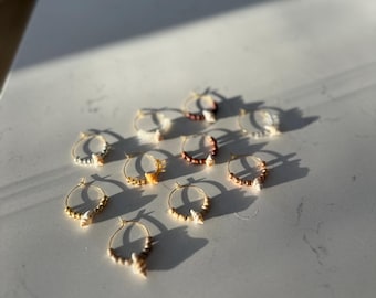 Könnte beinhalten: Eine Sammlung goldfarbener Creolen, jede mit kleinen dekorativen Elementen. Die Ohrringe sind auf einer weißen Oberfläche angeordnet, mit Schatten durch Sonnenlicht. Die Ohrringe haben ein Küsten-Thema.