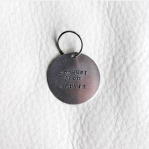 Op de afbeelding: Zilveren sleutelhanger met een rond label met de tekst "conquer from within".