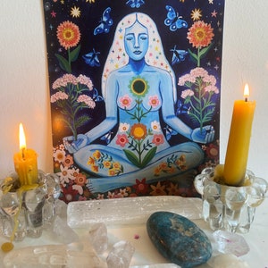 May include: A blue woman figure with flowers and stars on her body sits in a meditative pose. The background is a dark blue with stars and butterflies. The image is surrounded by candles, crystals, and flowers.