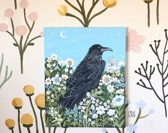May include: A painting of a black raven perched among white and blue flowers, set against a light blue sky with a crescent moon and stars. The artwork is on a square canvas, with a floral pattern on the wall behind it.