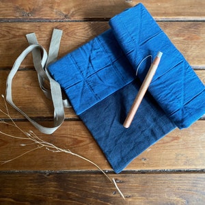 Op de afbeelding: Een blauwe stoffen rolhoes met een houten stylus erin. De hoes is gestikt en heeft een lichtbruin lint eromheen gebonden.