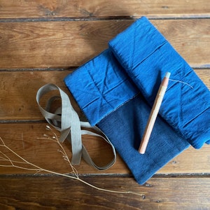 May include: A blue quilted fabric pouch with a wooden pencil on top of it. The pouch is lying on a wooden surface with a piece of beige ribbon next to it.
