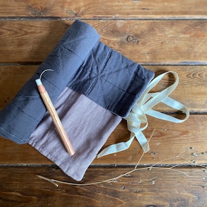 May include: A rolled-up grey and purple fabric pouch with a white ribbon. A wooden tool with a copper tip is lying next to the pouch.