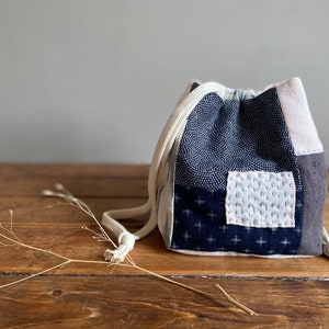 Könnte beinhalten: Eine kleine, blaue und weiße Kordeltasche aus Patchworkstoff. Die Tasche hat eine weiße Kordel und steht auf einer Holzoberfläche.