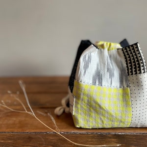 Pode incluir: Um par de sacos com cordão com um design de patchwork. Os sacos apresentam uma mistura de padrões, incluindo formas geométricas em preto e branco, listras verticais e um padrão xadrez amarelo brilhante. Os sacos são feitos de tecido e estão sobre uma superfície de madeira.