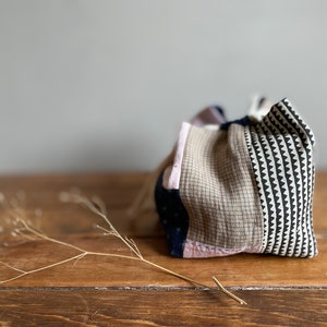 Pode incluir: Uma bolsa de tecido patchwork com fecho de cordão. A bolsa é feita de diferentes tecidos em tons de rosa, castanho, preto e branco. A bolsa está sobre uma superfície de madeira.