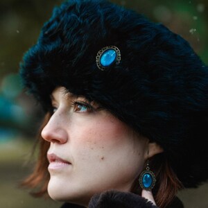 Könnte beinhalten: Eine Frau trägt einen schwarzen Pelz Hut mit einer blauen Edelstein Brosche und blauen Edelstein Ohrringen. Sie trägt eine dunkelblaue Jacke mit einem Pelzkragen.