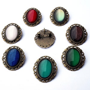 Può includere: Set di dieci spille ovali con pietre di vetro di diversi colori. Le spille sono in metallo con finitura oro antico. Le pietre sono incastonate in una montatura a castone e sono circondate da un bordo decorativo.