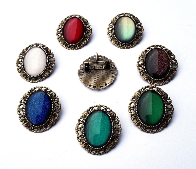 May include: Set of ten oval-shaped brooches with different colored glass stones. The brooches are made of metal with an antique gold finish. The stones are set in a bezel setting and are surrounded by a decorative border.
