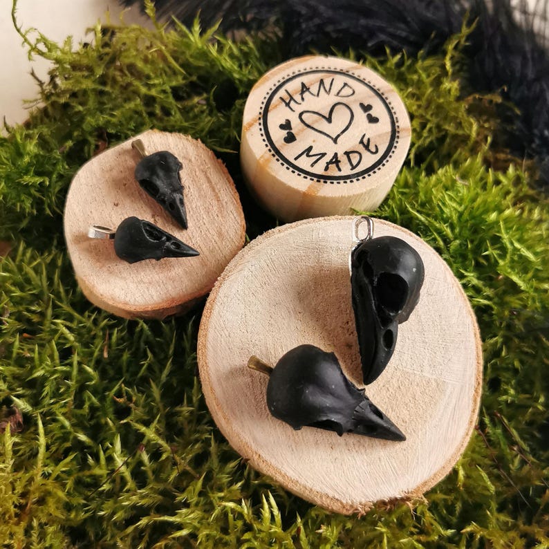 May include: Three black bird skull pendants on wooden bases. The pendants are on display on a bed of green moss. The wooden bases have the words "Hand Made" written on them.
