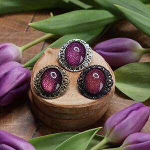 May include: Three oval-shaped brooches with ornate silver, bronze, and black metal frames. Each brooch features a sparkly, iridescent purple cabochon.