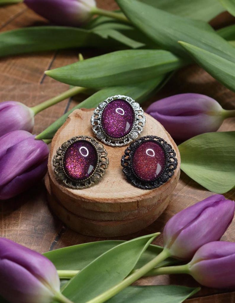 May include: Three oval-shaped brooches with ornate silver, bronze, and black metal frames. Each brooch features a sparkly, iridescent purple cabochon.
