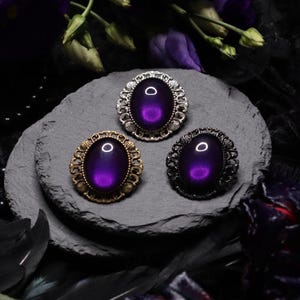 May include: Three oval-shaped brooches with ornate silver, gold, and black frames. Each brooch features a dark purple iridescent glass cabochon.