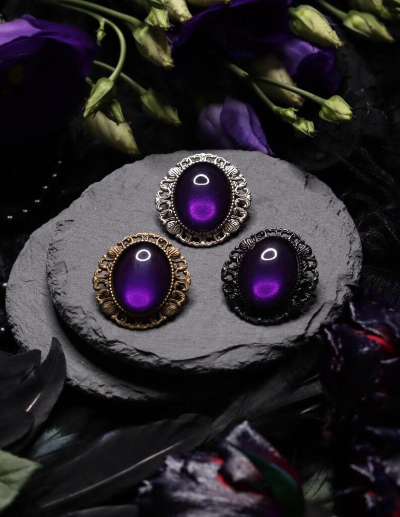 May include: Three oval-shaped brooches with ornate silver, gold, and black frames. Each brooch features a dark purple iridescent glass cabochon.