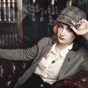 May include: A woman wearing a gray tweed blazer with a white button-down shirt underneath. She is wearing a brown and gray plaid newsboy cap and a gold necklace with a pendant. She is sitting on a brown leather couch with a bookshelf in the background.
