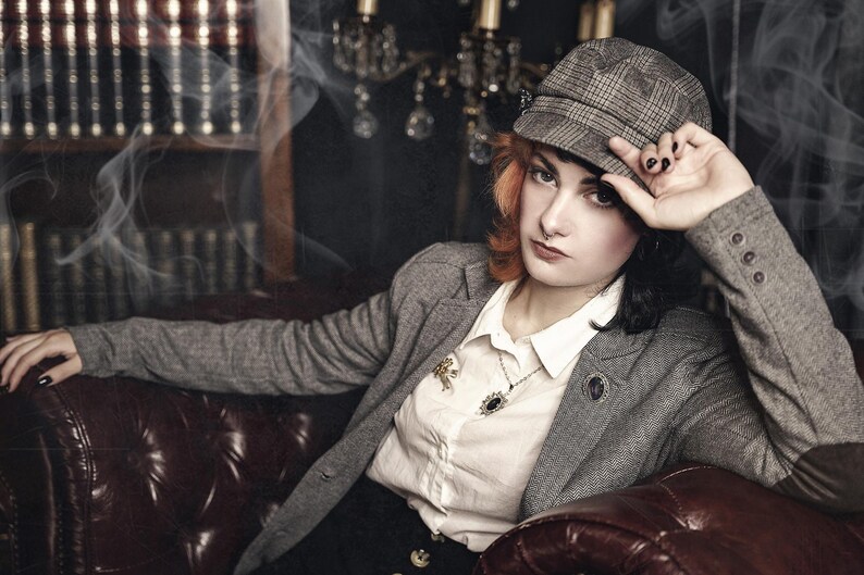 May include: A woman wearing a gray tweed blazer with a white button-down shirt underneath. She is wearing a brown and gray plaid newsboy cap and a gold necklace with a pendant. She is sitting on a brown leather couch with a bookshelf in the background.