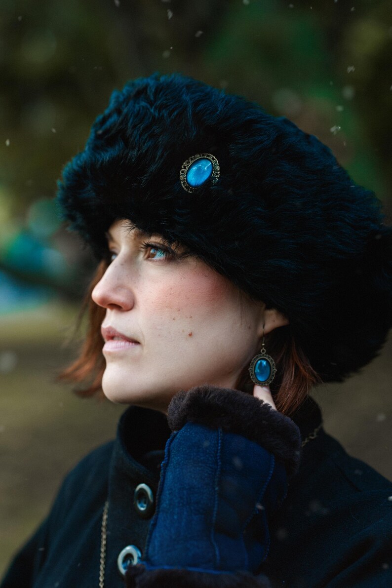 Könnte beinhalten: Eine Frau trägt einen schwarzen Pelz Hut mit einer blauen Edelstein Brosche und blauen Edelstein Ohrringen. Sie trägt eine dunkelblaue Jacke mit einem Pelzkragen.