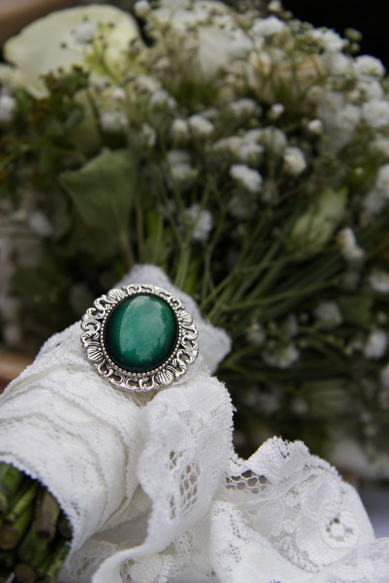 May include: A white lace ribbon tied around a bouquet of white baby's breath flowers with a silver ring featuring a green gemstone.
