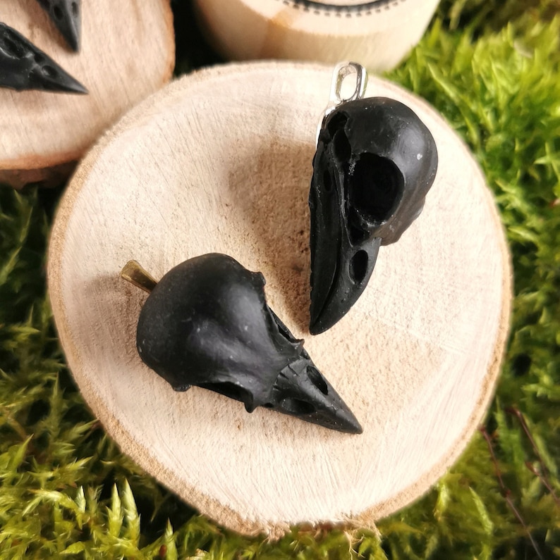 Könnte beinhalten: Zwei schwarze Rabenschädel-Anhänger auf einer Holzoberfläche. Die Schädel sind detailliert und haben eine matte Oberfläche.