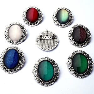 May include: Eight silver oval brooches with different colored glass stones. The stones are iridescent and change color in the light. The brooches are ornate and have a vintage style.